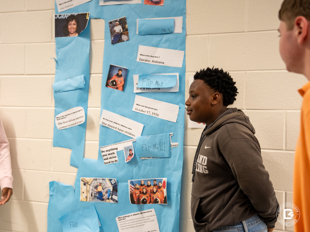 A student stands beside a blue poster display highlighting an African American astronaut, reading facts and looking at photos attached to the board.