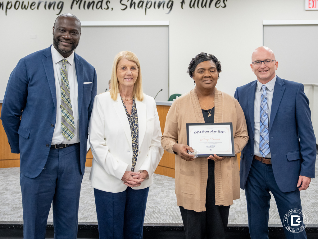 Four adults stand together at a Dorchester School District Four Board meeting. Mary Primus, Teacher at Woodland High School, holds a framed “DD4 Everyday Hero” certificate and a small award. District leaders stand beside her in the boardroom.