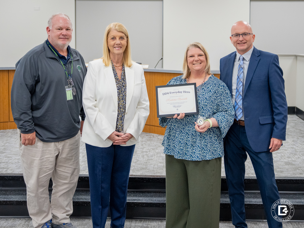 Four adults stand together at a Dorchester School District Four Board meeting. Kristina Brandt, School Counselor at Woodland Middle School, holds a framed “DD4 Everyday Hero” certificate and a small award. She stands with district leaders in a boardroom setting.
