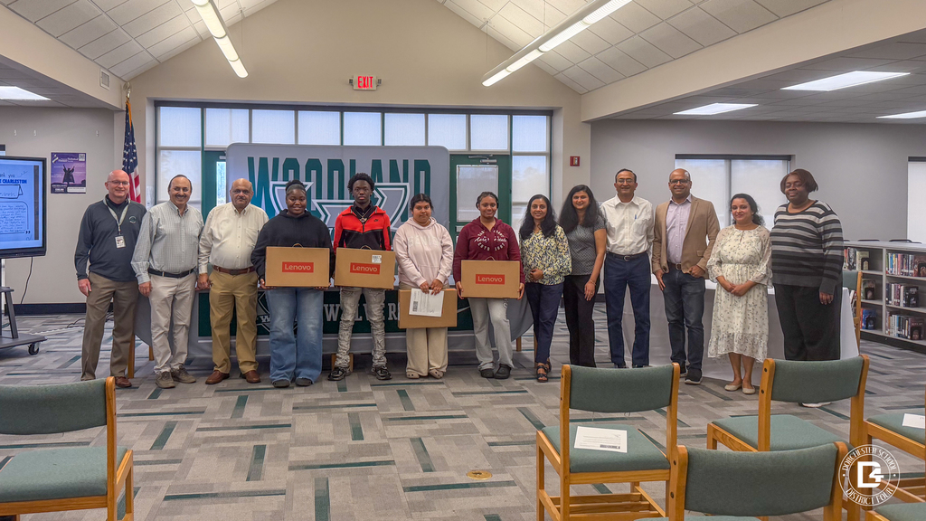 Four Woodland High School seniors stand in front of a large Woodland Wolverines backdrop holding boxed Lenovo laptops, joined by Namaste Charleston representatives and school staff for a group photo in the media center.