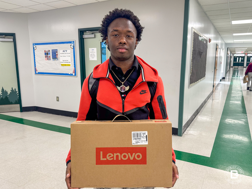 A Woodland High School senior stands in a hallway holding a boxed Lenovo laptop and smiling, with school office doors and vending machines visible in the background.