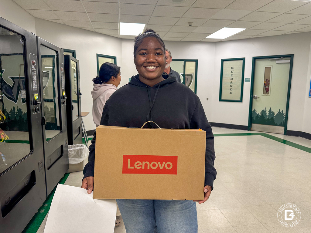 A Woodland High School senior wearing a red and black jacket holds a boxed Lenovo laptop in a school hallway.