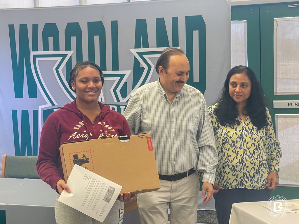 A Woodland High School senior receives a boxed Lenovo laptop from a Namaste Charleston representative in front of a Woodland Wolverines backdrop.