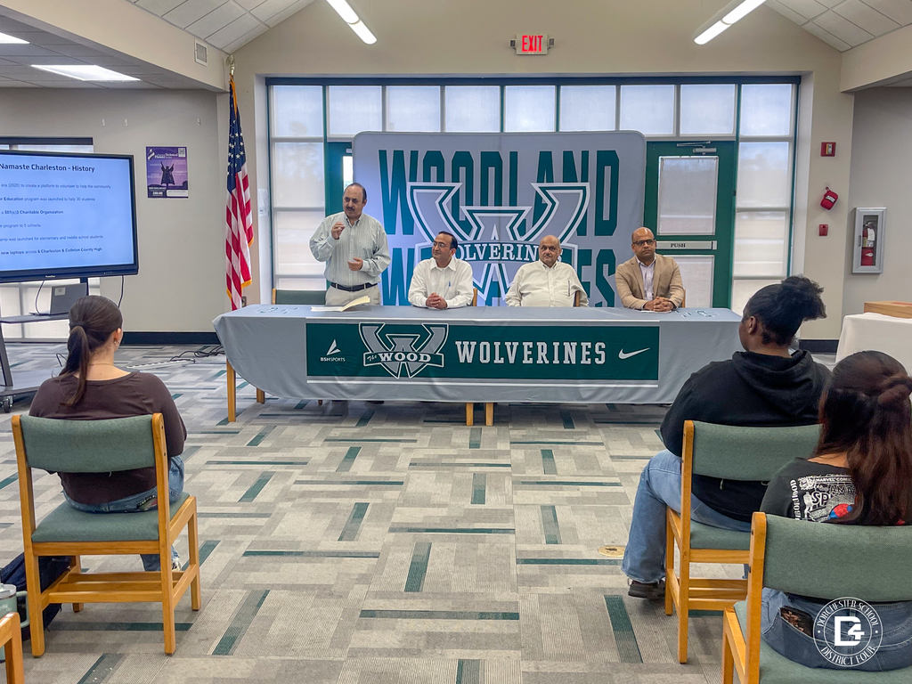 Namaste Charleston representatives sit at a table with a Woodland Wolverines banner while one presenter gestures toward a screen displaying information about the organization’s history.