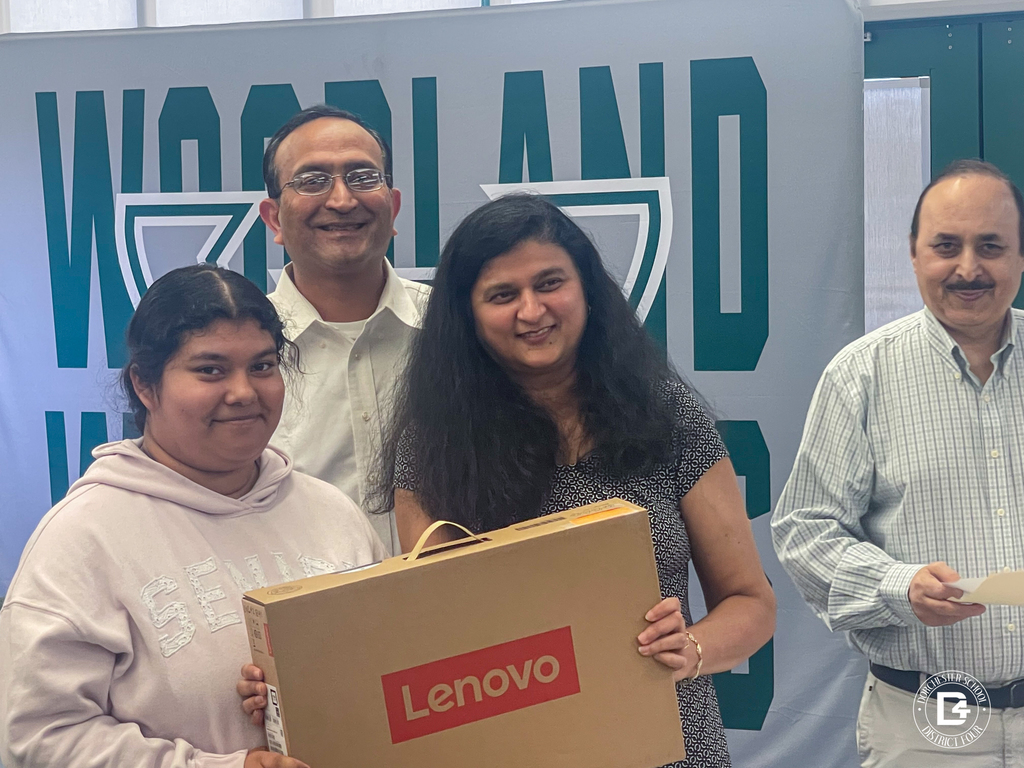 A Woodland High School senior holds a boxed Lenovo laptop and a document while standing beside two Namaste Charleston representatives in front of the Woodland Wolverines backdrop.