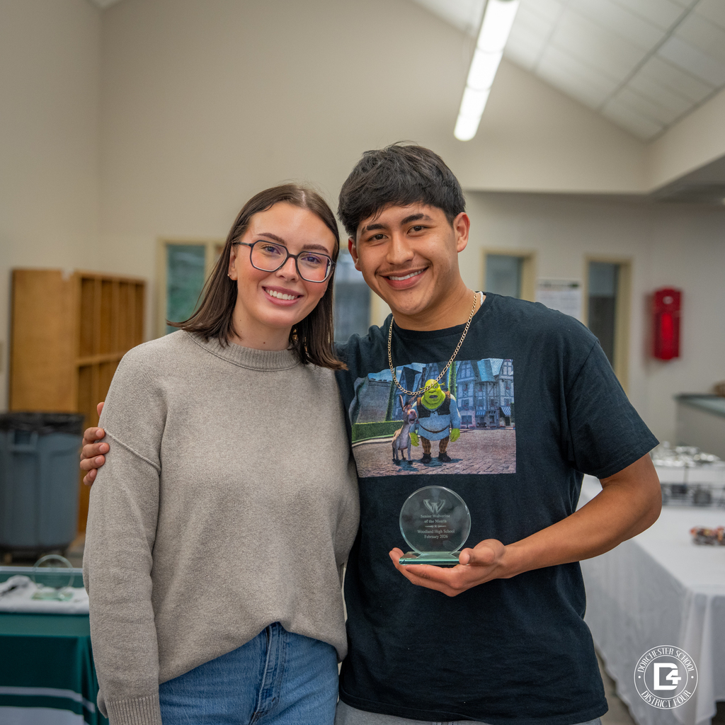 Female staff member and male student smile together in the media center as he holds his glass Wolverine of the Month award.