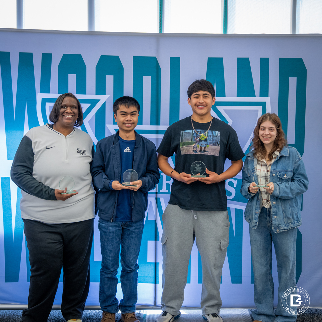 Two staff members and two students stand in front of the Woodland Wolverines backdrop holding their glass Wolverine of the Month awards.