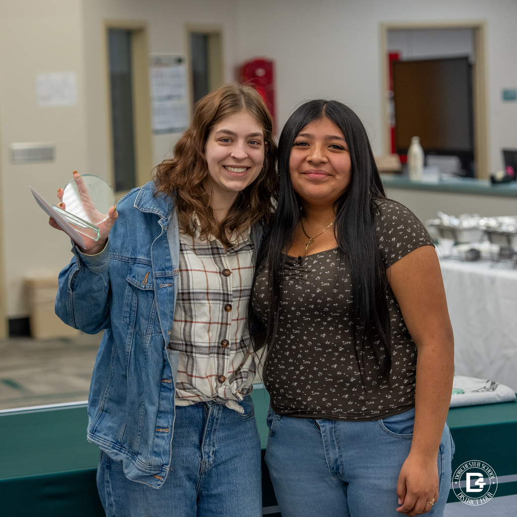 Female  staff member stand next to a female student as they smile together, one holding her glass Wolverine of the Month award.
