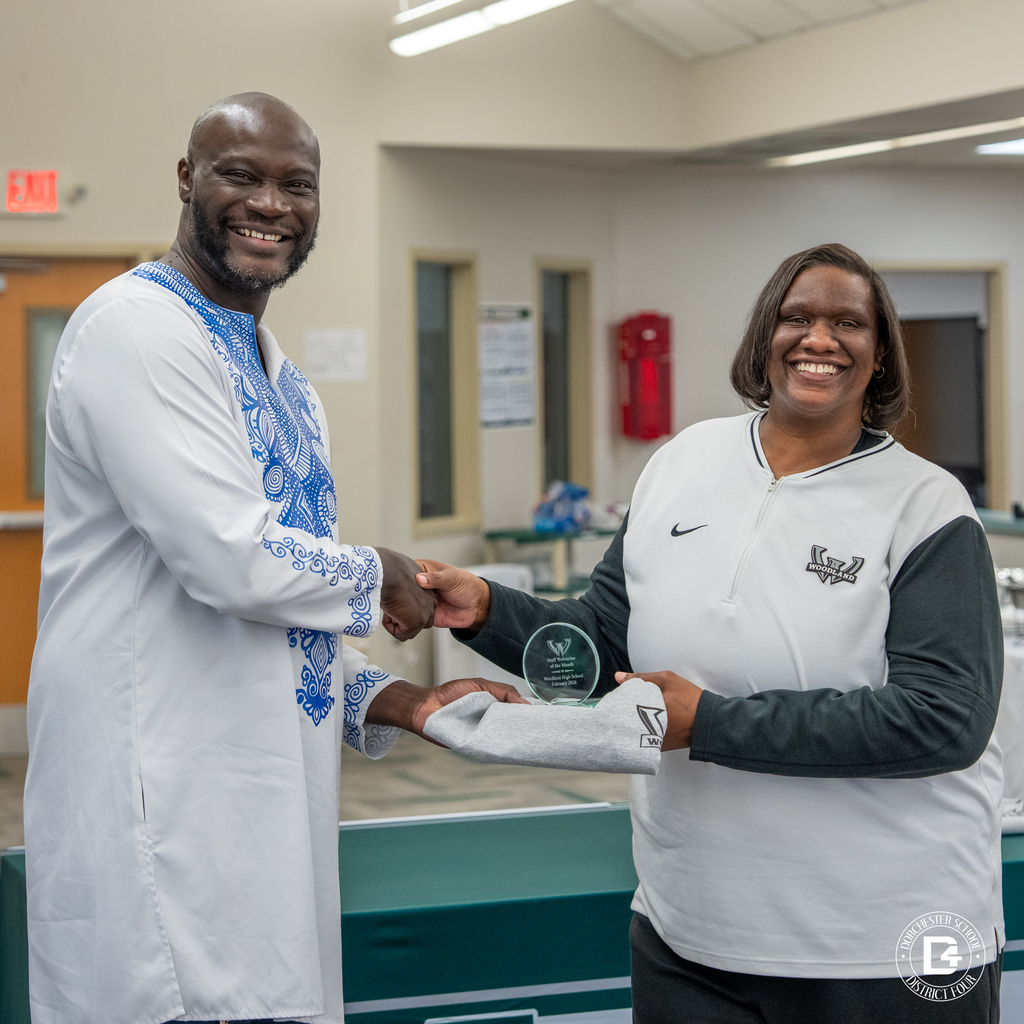 Administrator shakes hands with a female staff member recognized as a Wolverine of the Month while she holds her award and Woodland shirt.