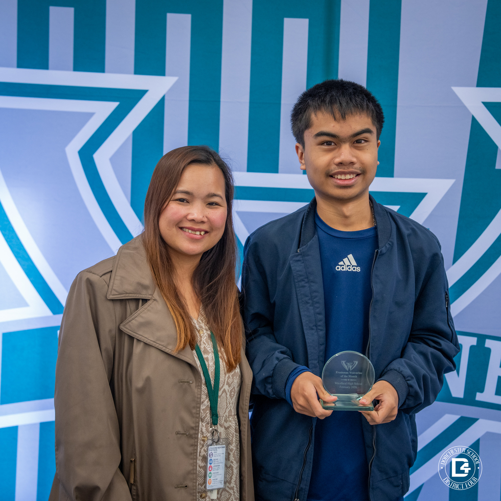 Female staff member stands beside a male student holding his Wolverine of the Month award in front of the Woodland backdrop.