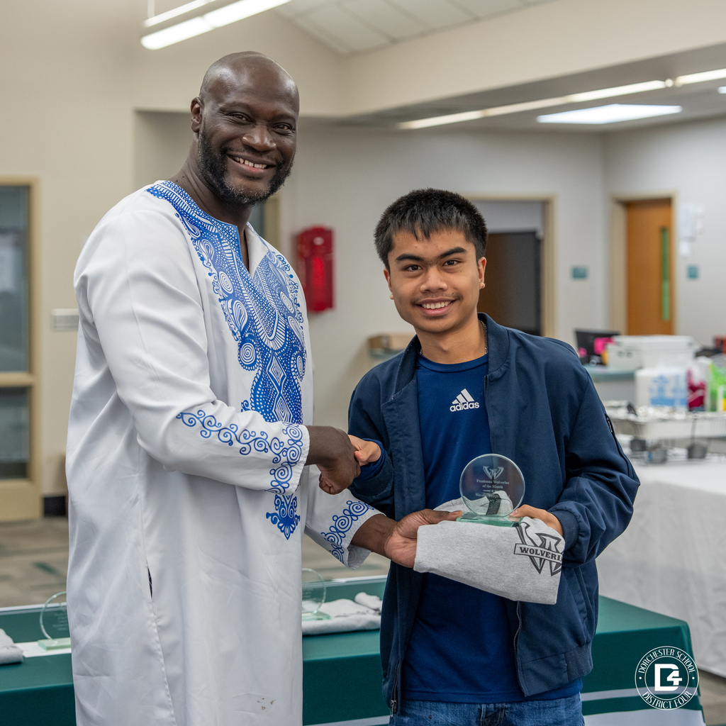 Administrator shakes hands with a male student receiving a glass Wolverine of the Month award and Woodland shirt in the media center