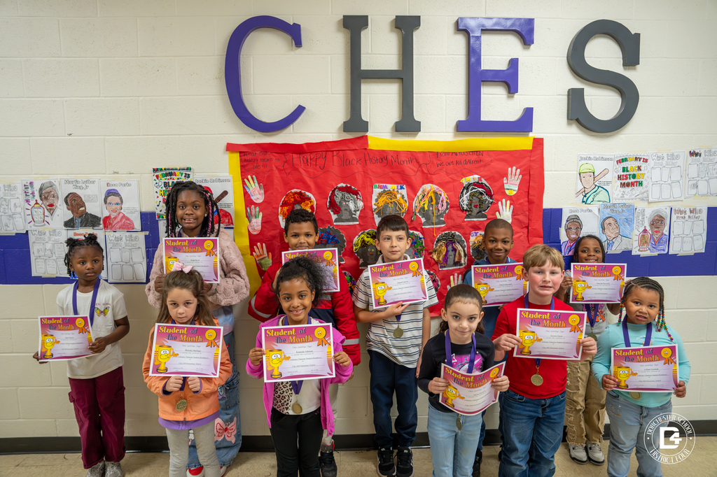 A group of elementary students stand in a school hallway under large “CHES” letters, holding “Student of the Month” certificates and wearing medals. Behind them is a red “Happy Black History Month” display board with student artwork and portraits. The students were recognized at Clay Hill Elementary School for demonstrating kindness.