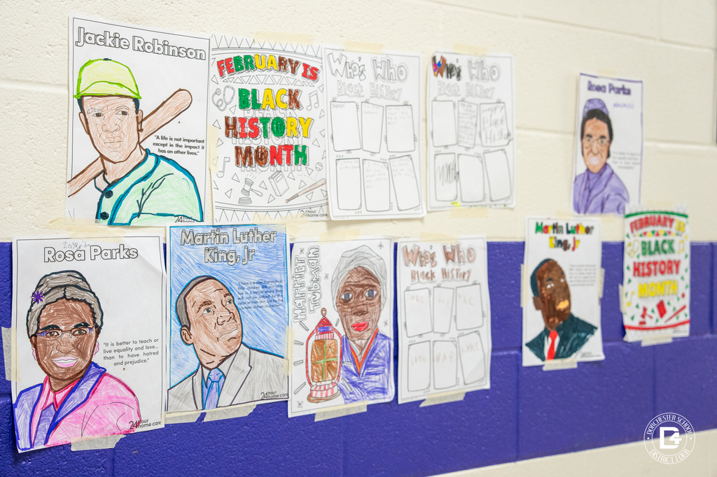 A hallway wall at Clay Hill Elementary displays student Black History Month projects, including colored portraits of Martin Luther King Jr., Rosa Parks, Jackie Robinson, and “Who’s Who in Black History” worksheets taped along a blue stripe on a white cinderblock wall.