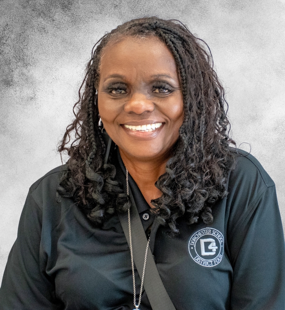 Portrait of Mrs. Anntionette Haynes smiling at the camera. She is wearing a black Dorchester School District Four polo shirt with the district logo on the left chest and a gray background behind her.