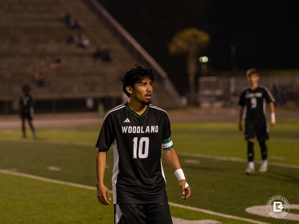A Woodland boys soccer player wearing number 10 walks across the field during a night game with teammates visible in the background.