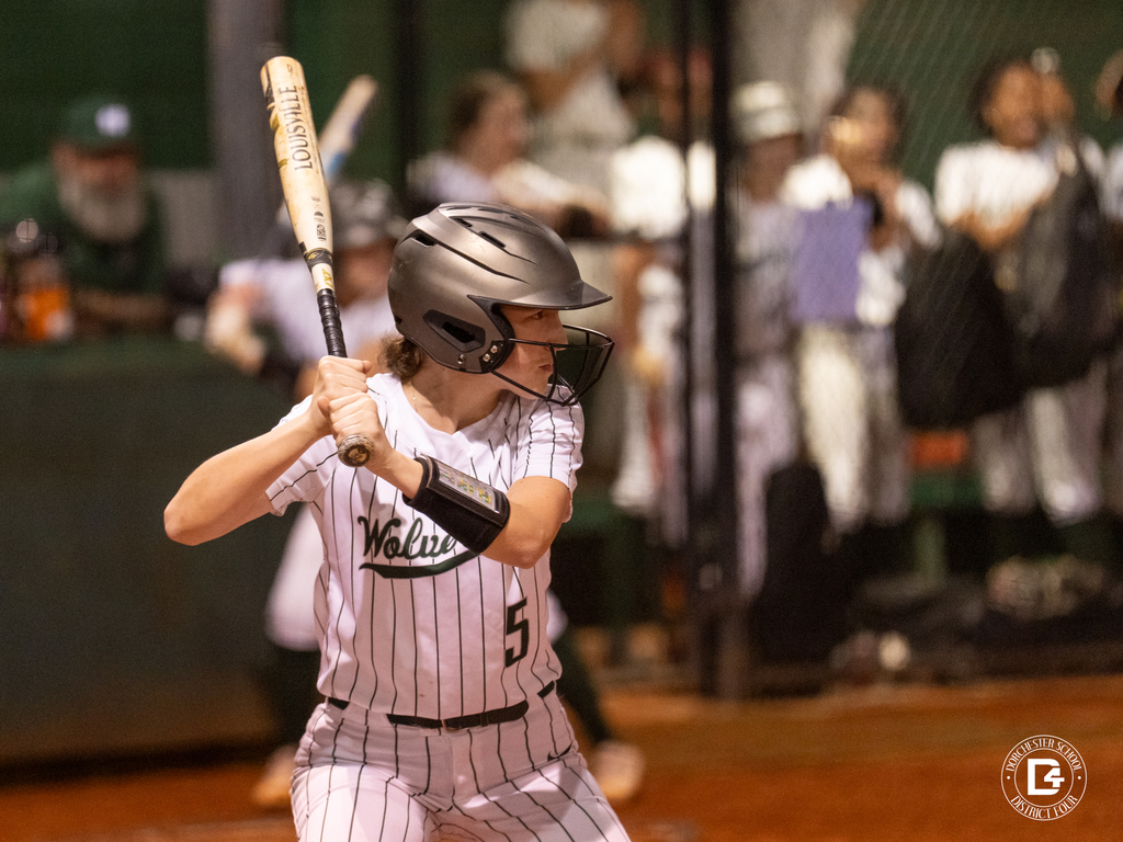 A Woodland softball player wearing number 5 prepares to bat while teammates and spectators watch from the dugout behind her.
