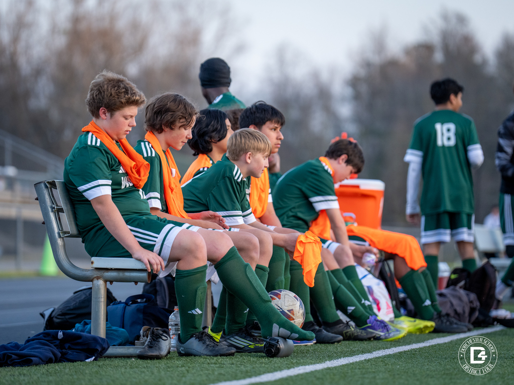 Woodland soccer players sit on the bench during the match wearing green uniforms and orange training vests while watching the action on the field.