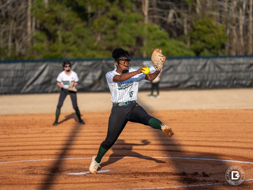 A Woodland softball pitcher in a white Wolverines uniform releases a pitch during a game while an infielder prepares behind her on the dirt field.