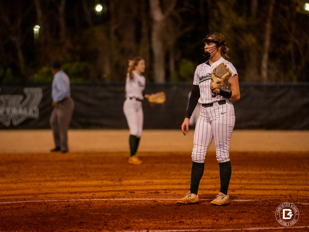 A Woodland softball infielder stands ready in the field during a night game while another player and an umpire are visible in the background.