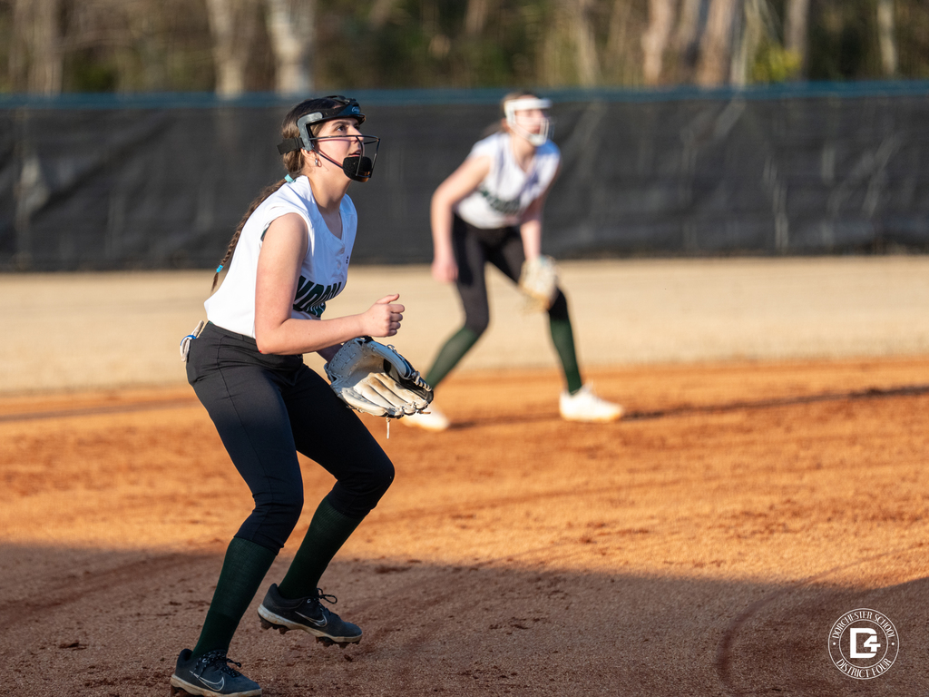 A Woodland softball player wearing a face guard and glove watches a ball in the air while another infielder stands ready behind her on the infield.