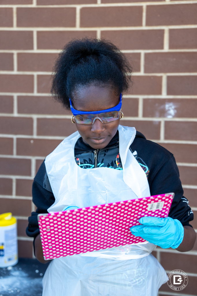 Student wearing safety goggles, gloves, and a plastic apron records observations on a clipboard during Dino Dig Day in Mrs. Rhode’s 8th grade science class at Woodland Middle School.