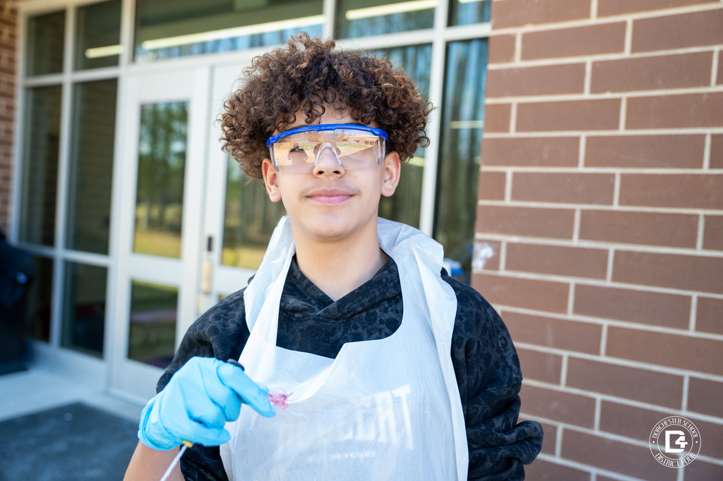 A Woodland Middle School student wearing goggles and a protective apron holds a small fossil discovery during the Dino Dig Day science experiment.