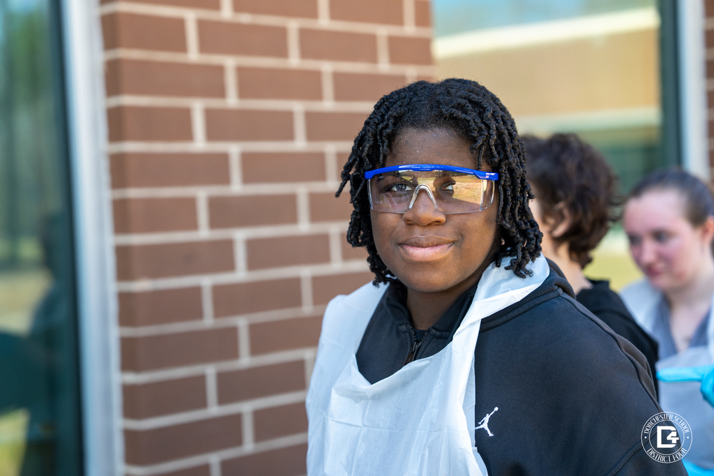 A student wearing safety goggles and gloves pauses while participating in the hands-on fossil excavation activity during Dino Dig Day.