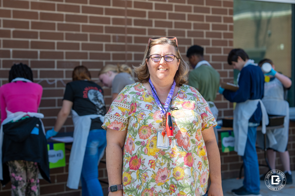 Mrs. Rhode stands in front of her class while students work behind her chipping and examining materials during the Dino Dig Day fossil investigation.