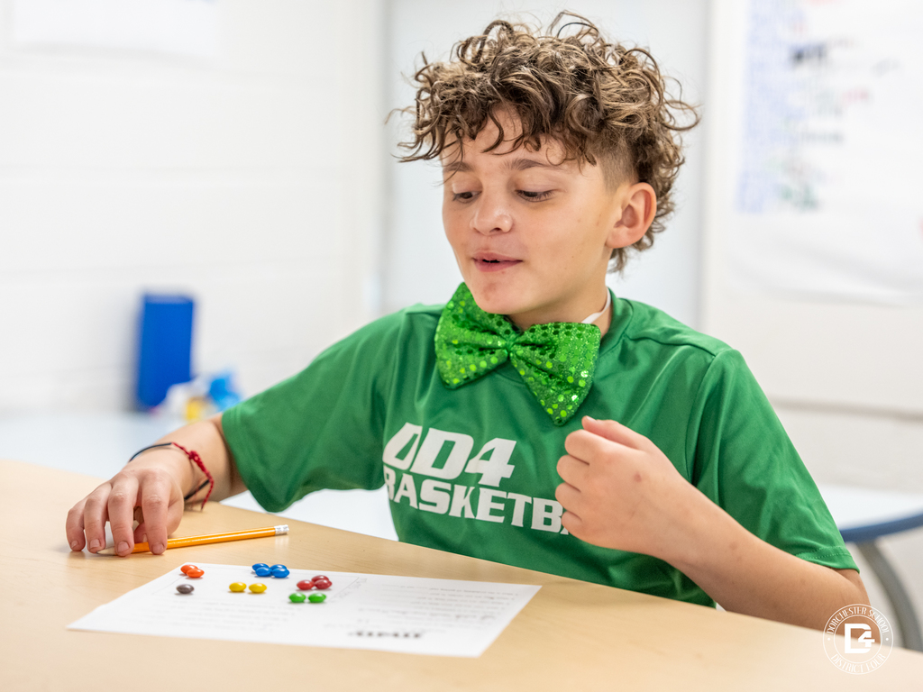 Student wearing a green shirt and bow tie reacts excitedly while working on a probability activity using M&M’s.
