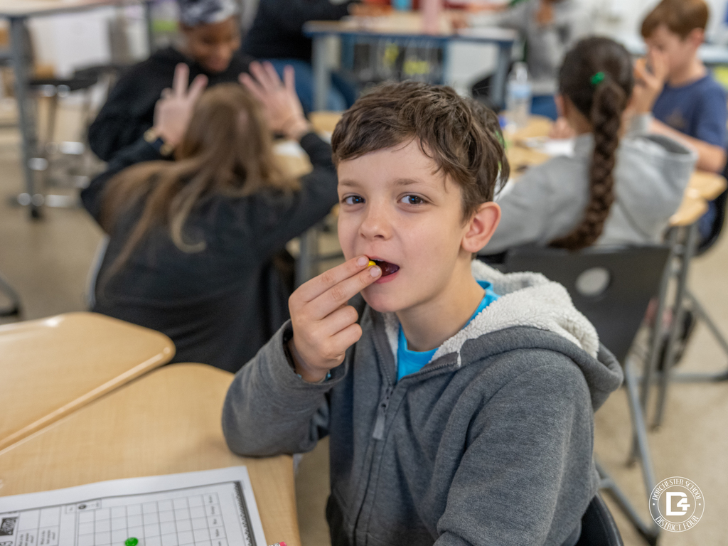 Student places an M&M in their mouth while seated at a desk during the hands-on math lesson.