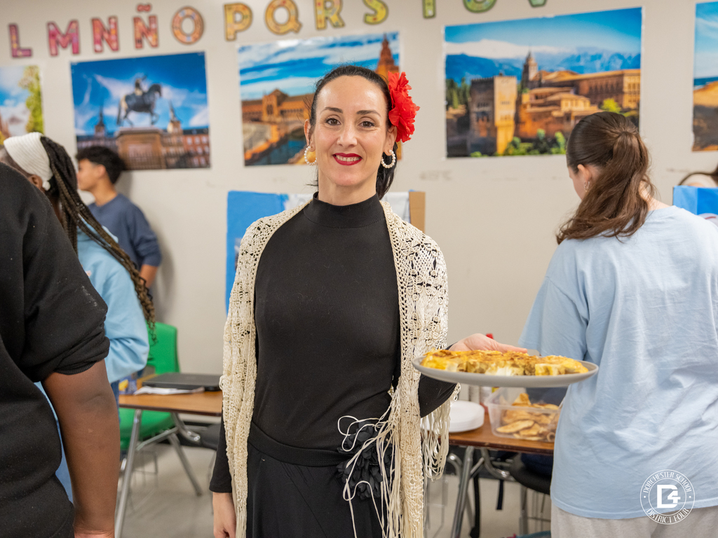 Teacher stands in the classroom holding a plate of traditional food while students present their country projects during Spanish class.