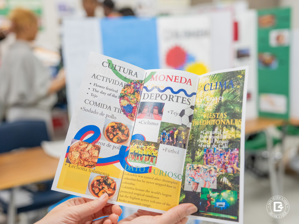 Hands hold a colorful informational brochure about Colombian culture that includes sections on activities, food, sports, climate, and traditional festivals.
