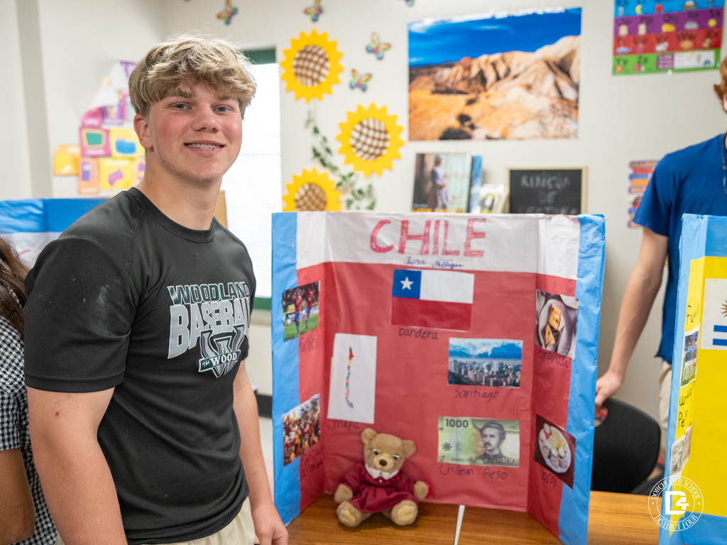 Student stands next to a presentation board about Chile displaying the national flag, currency, map, and images of Santiago.