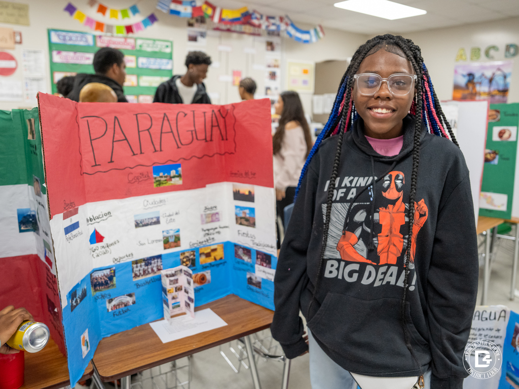 Student stands beside a presentation board about Paraguay that highlights cities, sports, food, and cultural facts.