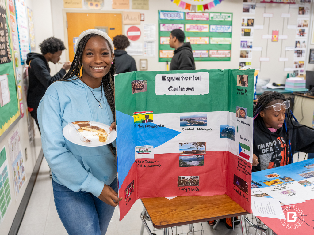 Student smiles while holding a plate of food next to a presentation board about Equatorial Guinea featuring photos, landmarks, and cultural information.