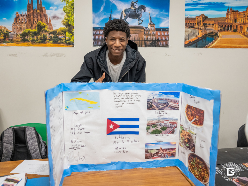 Student gives a thumbs-up behind a presentation board about Cuba featuring the national flag, cities, foods, and cultural details.