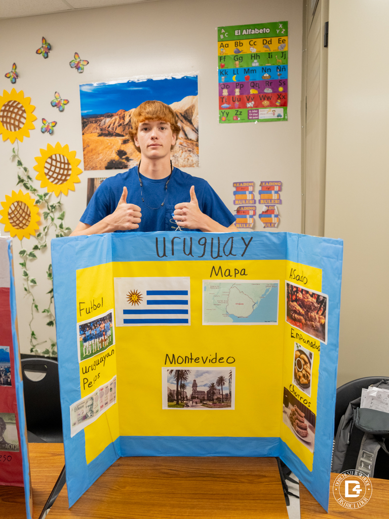 Student gives a thumbs-up while standing behind a presentation board about Uruguay that includes the flag, map, foods, and facts about Montevideo.