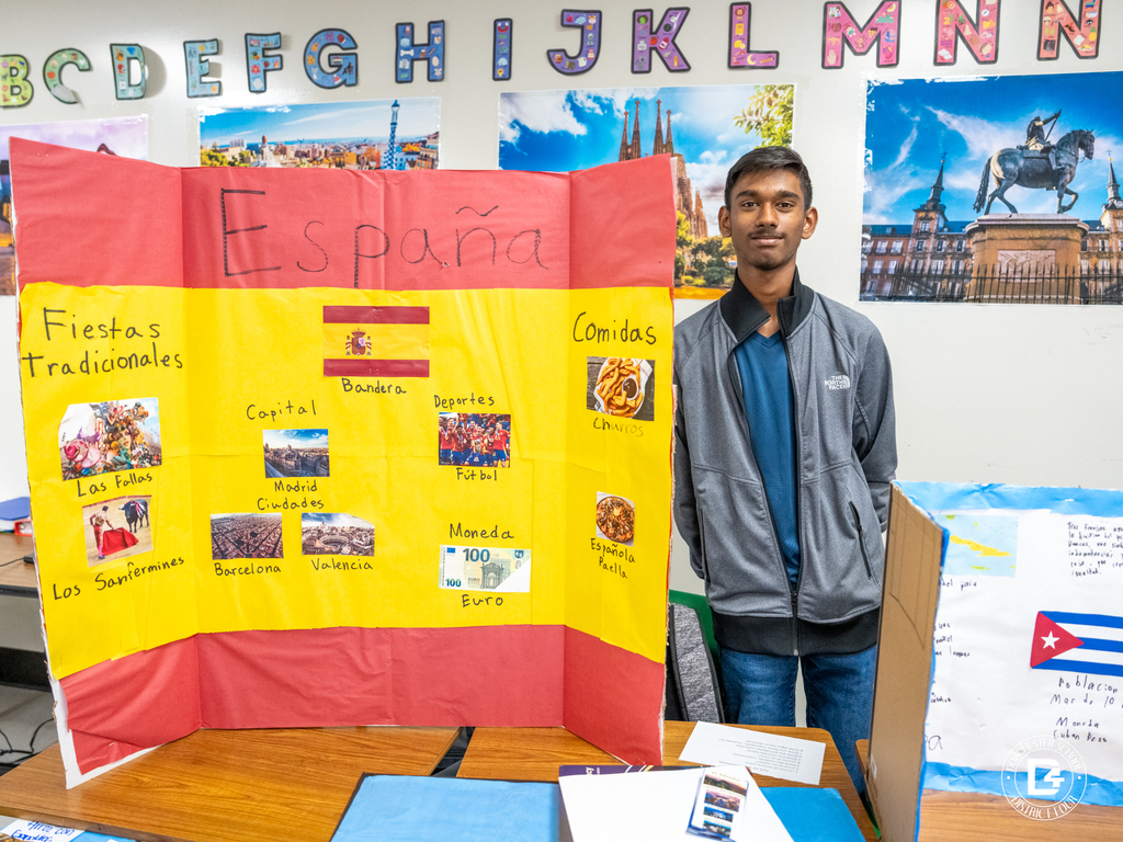 Student stands beside a presentation board about Spain that includes the Spanish flag, cities, traditional festivals, foods, sports, and currency.