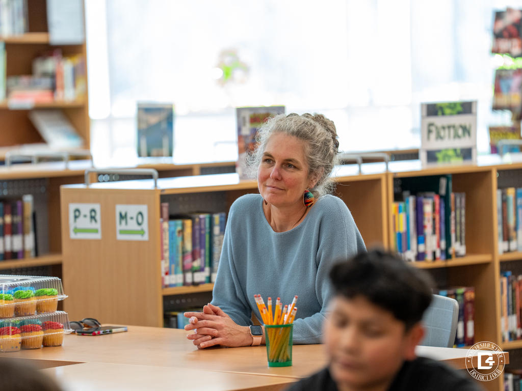 Mrs. Weather sits at a table listening while students participate in the leadership presentation.