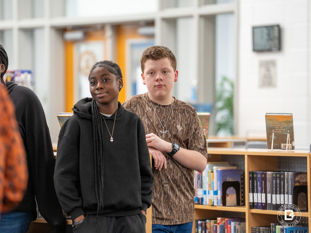 Two students stand near the bookshelves listening to the speaker during the ProTeam leadership session in the Woodland Middle School library.