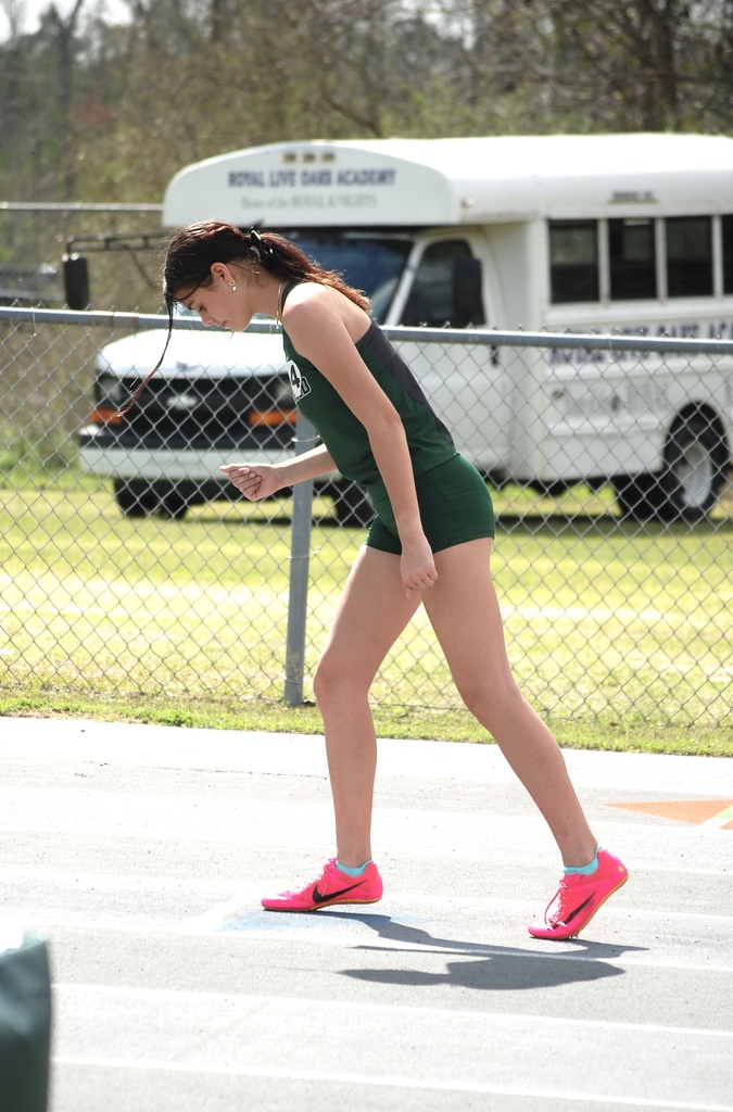 Woodland High School athlete walks along the track preparing for her event while a bus and fencing appear in the background.