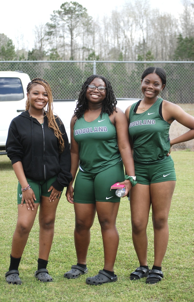 Three Woodland High School girls track athletes stand together and smile beside the field during the meet.