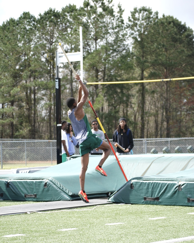 Woodland High School athlete clears the bar during a pole vault attempt while teammates watch from near the landing mat.