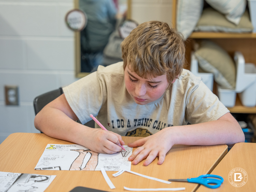 A student sits at a desk concentrating while writing and coloring on a character analysis worksheet about Nya from A Long Walk to Water. Scissors and paper strips sit beside the project.