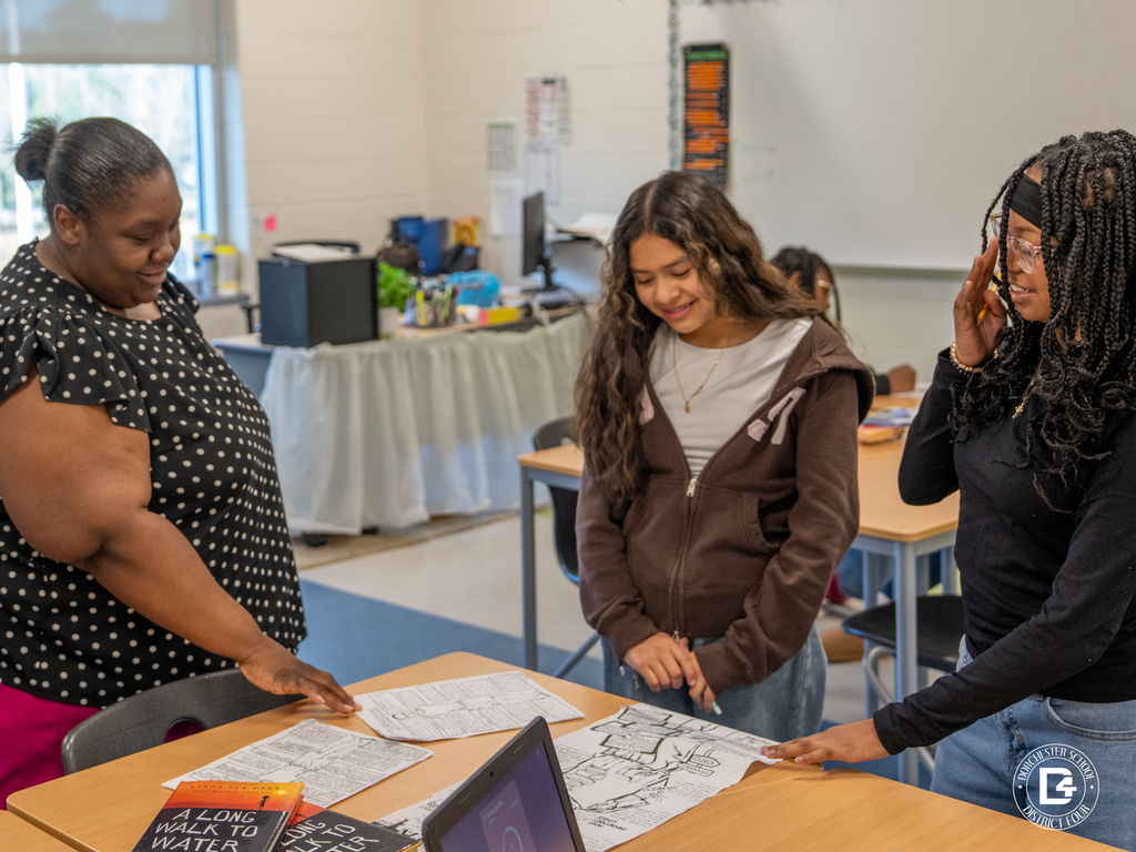 Ms. Smith stands with students at a table as they review and discuss their completed character analysis pages based on the novel A Long Walk to Water.