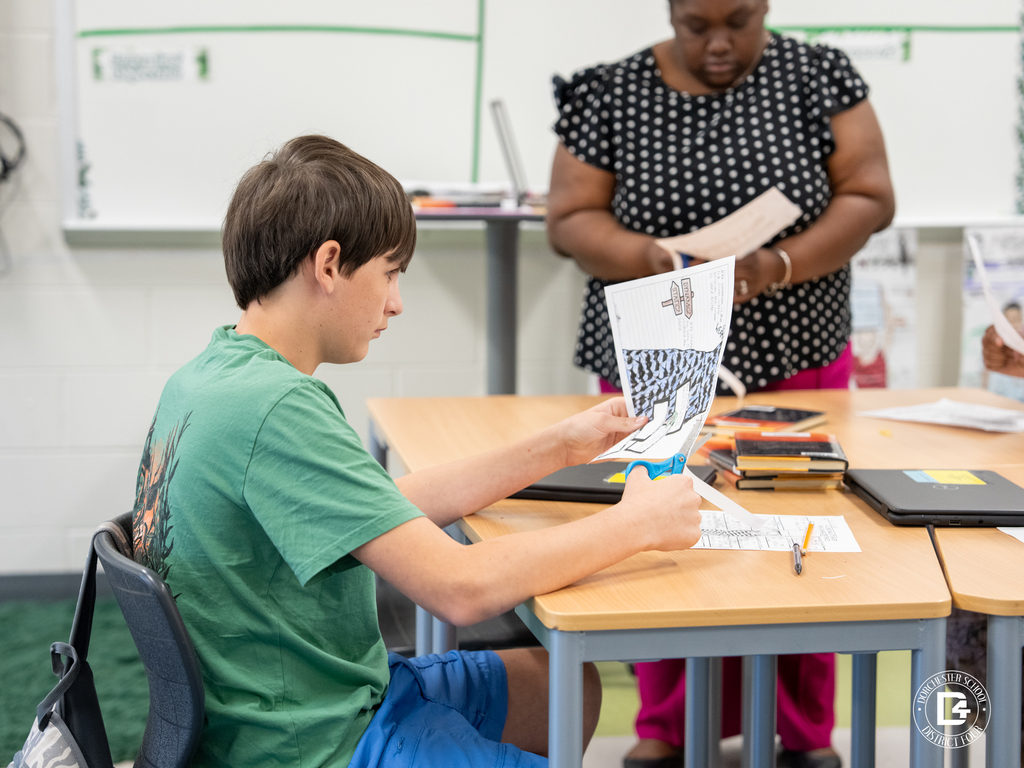 A student carefully cuts out sections of a character analysis worksheet while the teacher assists in the background.