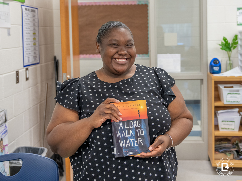 Teacher stands in the classroom smiling while holding the book A Long Walk to Water by Linda Sue Park, which students are studying for a character analysis activity.
