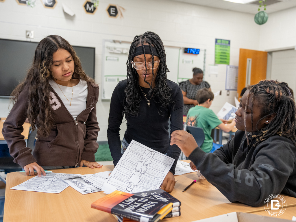 A group of students gather around a table comparing their character analysis work from A Long Walk to Water while discussing the story.