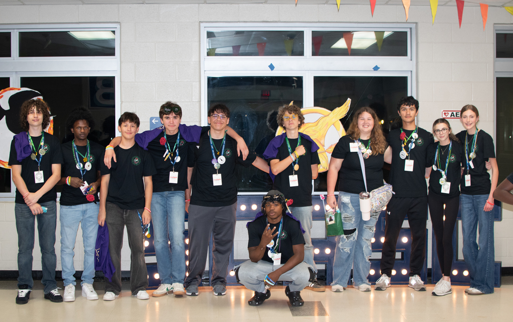 Woodland High School Student Council members pose together inside Stall High School during a leadership conference, wearing black Student Council shirts and conference badges while standing in front of a decorative backdrop with lights and banners, with one student kneeling in front of the group.