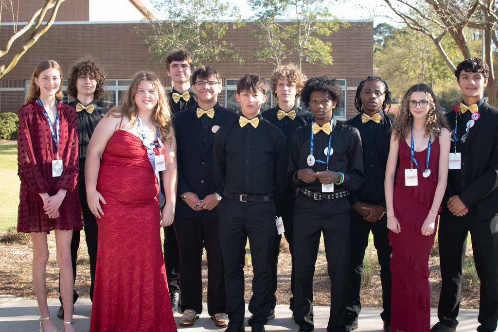 A group of Woodland High School Student Council members stand outside in formal attire, including black outfits with gold bow ties and red dresses, wearing conference badges and medals while posing together in front of a school building during the leadership conference at Stall High School.